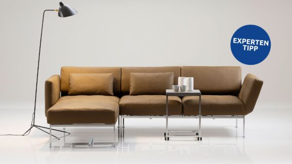 Ein modernes, braunes Sofaprogramm „roro“ von Brühl mit einer Chaiselongue, einem Beistelltisch aus Metall, auf dem Tassen gestapelt sind, und einer schwarzen Stehlampe vor einem schlichten weißen Hintergrund. An der Wand befindet sich ein blauer „Expertentipp“-Aufkleber.