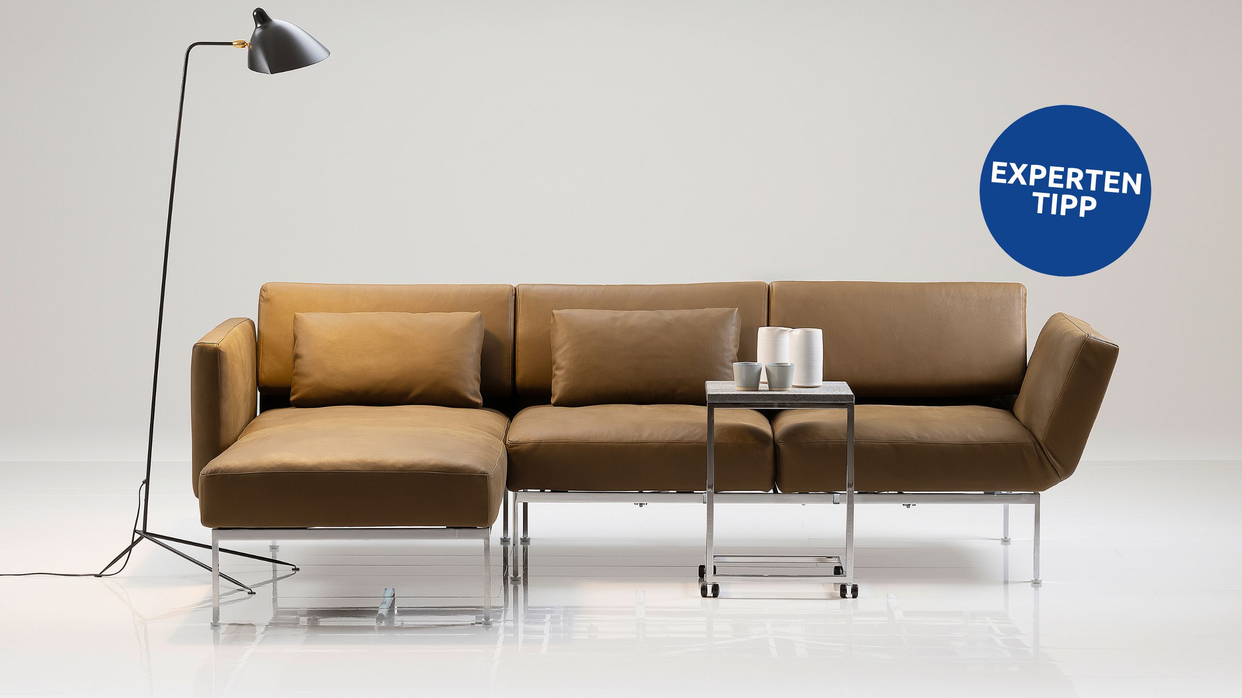Ein modernes, braunes Sofaprogramm „roro“ von Brühl mit einer Chaiselongue, einem Beistelltisch aus Metall, auf dem Tassen gestapelt sind, und einer schwarzen Stehlampe vor einem schlichten weißen Hintergrund. An der Wand befindet sich ein blauer „Expertentipp“-Aufkleber.