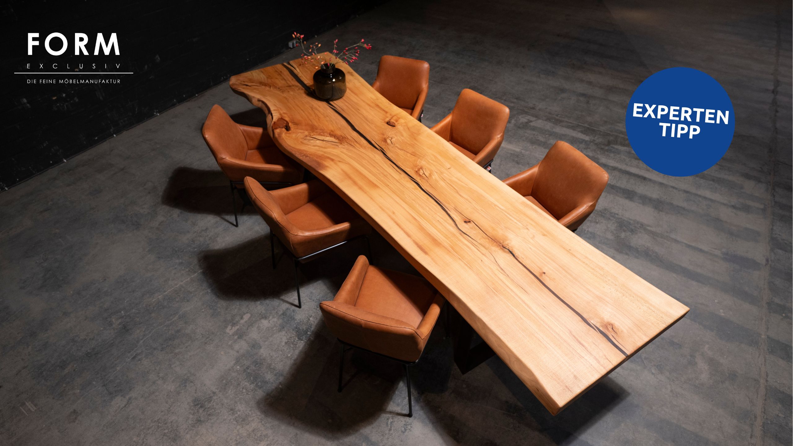 Großer Form Exclusiv Esstisch Monolith mit natürlicher Maserung, umgeben von acht braunen Lederstühlen auf einem Betonboden; eine schwarze Vase mit Zweigen steht auf dem Tisch, entworfen von Marcus Braum.