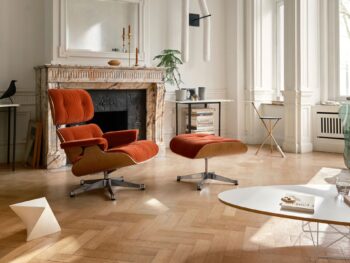 Ein modernes Wohnzimmer mit einem Marmorkamin, einem orangefarbenen Loungesessel und einer Ottomane, einem Schreibtisch, großen Fenstern und minimalistischem Dekor schafft einen gemütlichen Rückzugsort im Winter, inspiriert von Vitra Homestories und einem Gefühl von Harmonie.