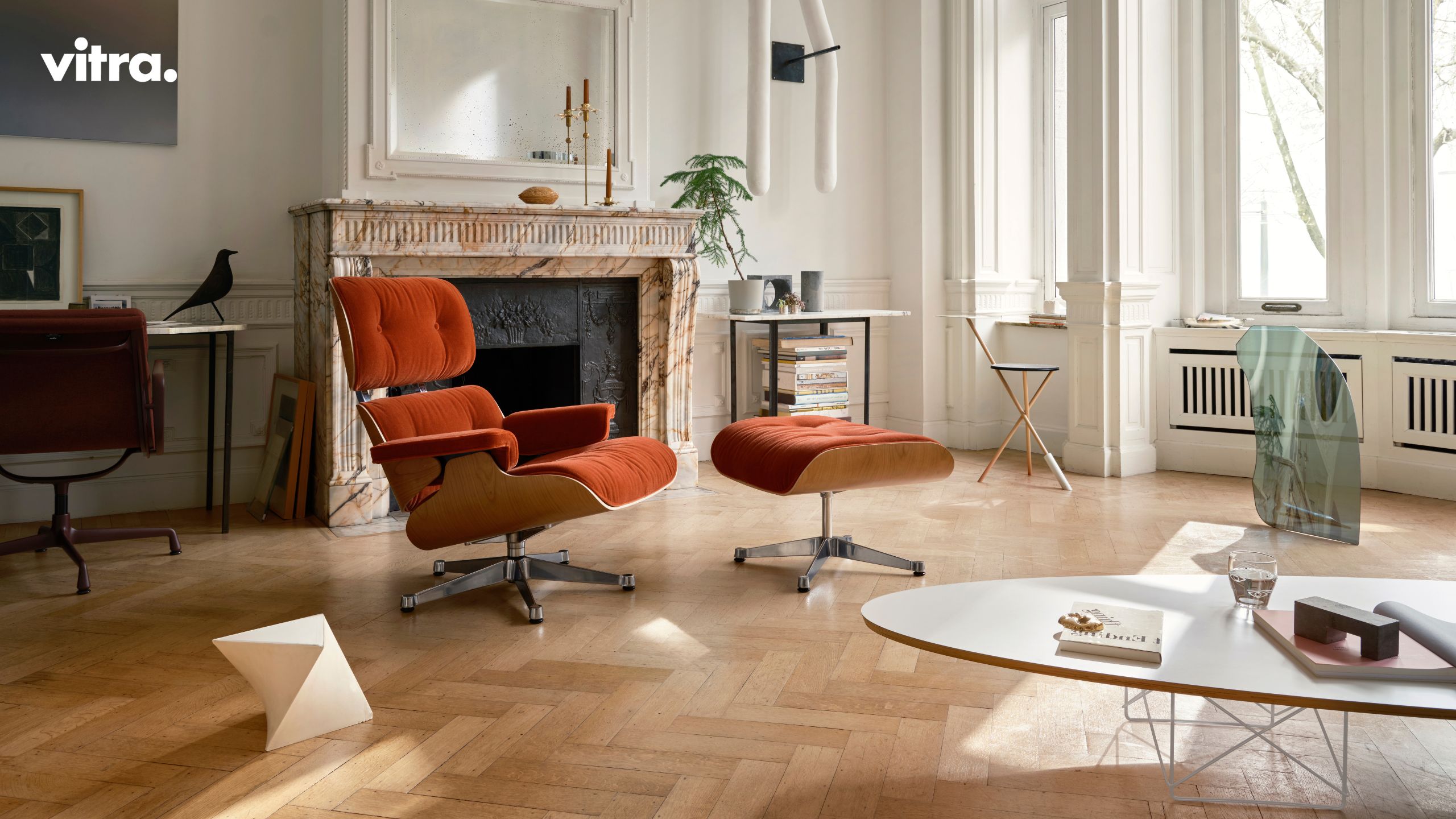 Ein modernes Wohnzimmer mit einem Marmorkamin, einem orangefarbenen Loungesessel und einer Ottomane, einem Schreibtisch, großen Fenstern und minimalistischem Dekor schafft einen gemütlichen Rückzugsort im Winter, inspiriert von Vitra Homestories und einem Gefühl von Harmonie.