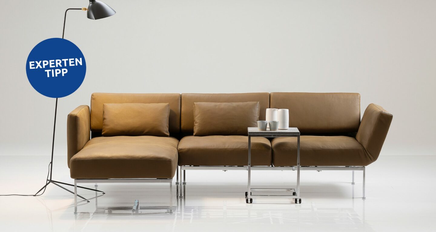 Ein braunes Ecksofa mit Metallbeinen, ein kleiner Beistelltisch aus Metall mit weißem Dekor und eine schwarze Stehlampe in einem minimalistischen, weißen Ambiente – Inspiration für modernen Stil vom Möbelhaus Bad Homburg. Links ein blauer Kreis mit der Aufschrift „EXPERTEN TIPP“. Ein braunes Ecksofa mit Metallbeinen, ein kleiner Beistelltisch aus Metall mit weißem Dekor und eine schwarze Stehlampe in einem minimalistischen, weißen Ambiente – Inspiration für modernen Stil vom Möbelhaus Bad Homburg. Links ein blauer Kreis mit der Aufschrift „EXPERTEN TIPP“.