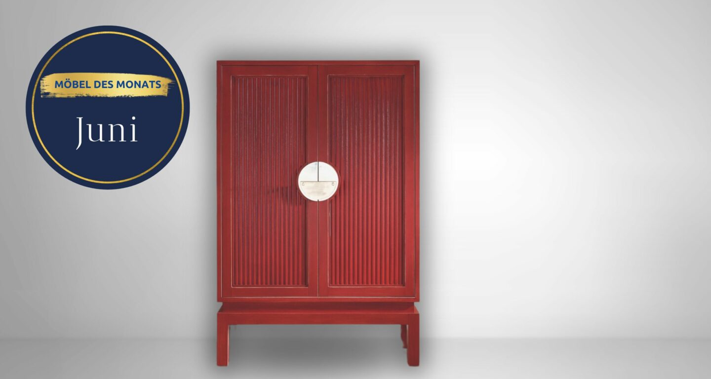 Ein roter Holzschrank mit vertikalen Rillen und einem runden Metallgriff steht vor einem schlichten hellgrauen Hintergrund. Auf einem blau-goldenen Schild steht „Möbel des Monats Juni“ vom Möbelhaus Bad Homburg.
