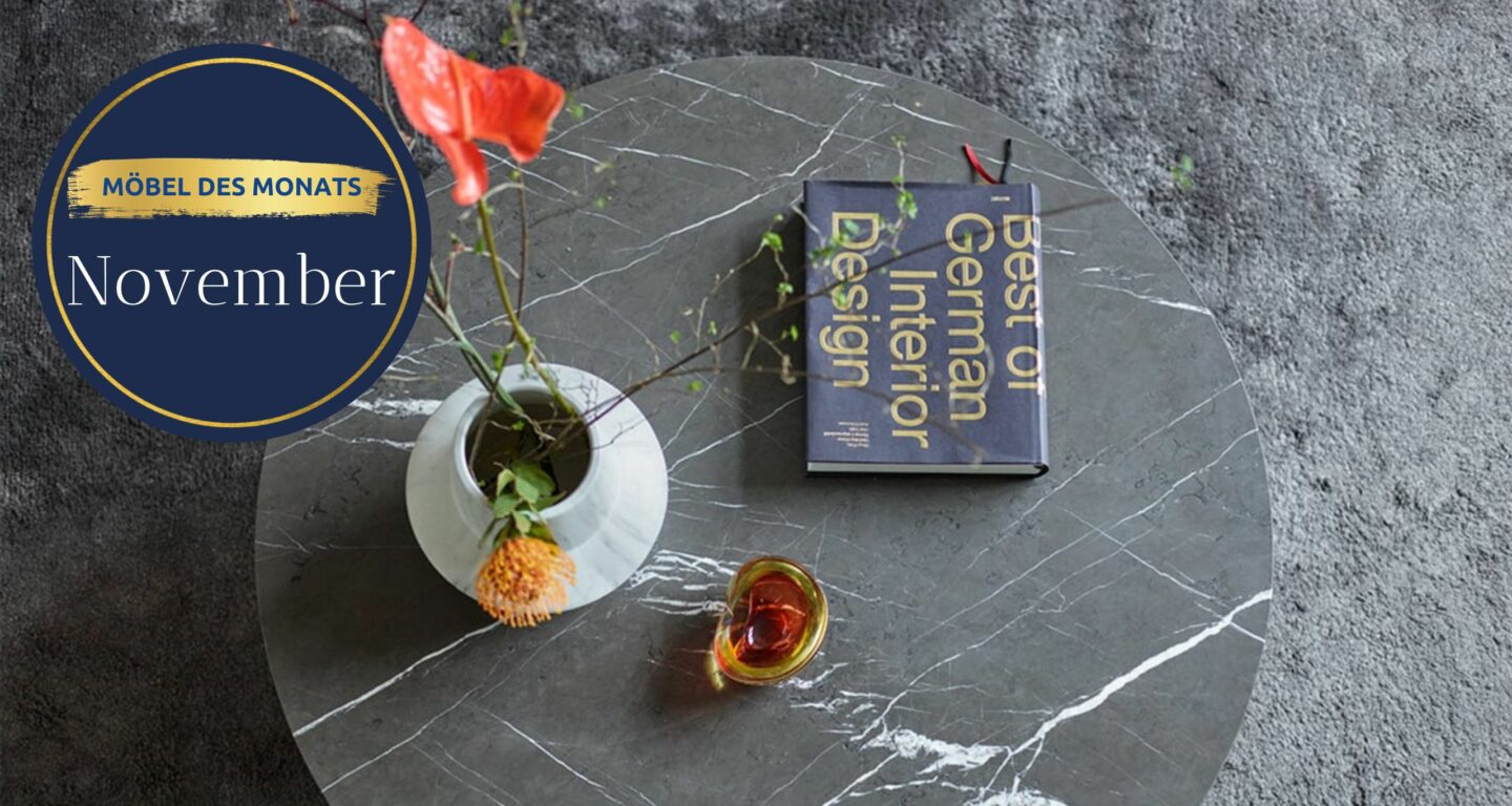 Ein runder grauer Marmortisch vom Möbelhaus Bad Homburg mit einer weißen Blumenvase, einem Orangendrink und dem Buch Best of German Interior Design steht auf einem grauen Teppich. Auf dem Schild steht Ein runder grauer Marmortisch vom Möbelhaus Bad Homburg mit einer weißen Blumenvase, einem Orangendrink und dem Buch Best of German Interior Design steht auf einem grauen Teppich. Auf dem Schild steht