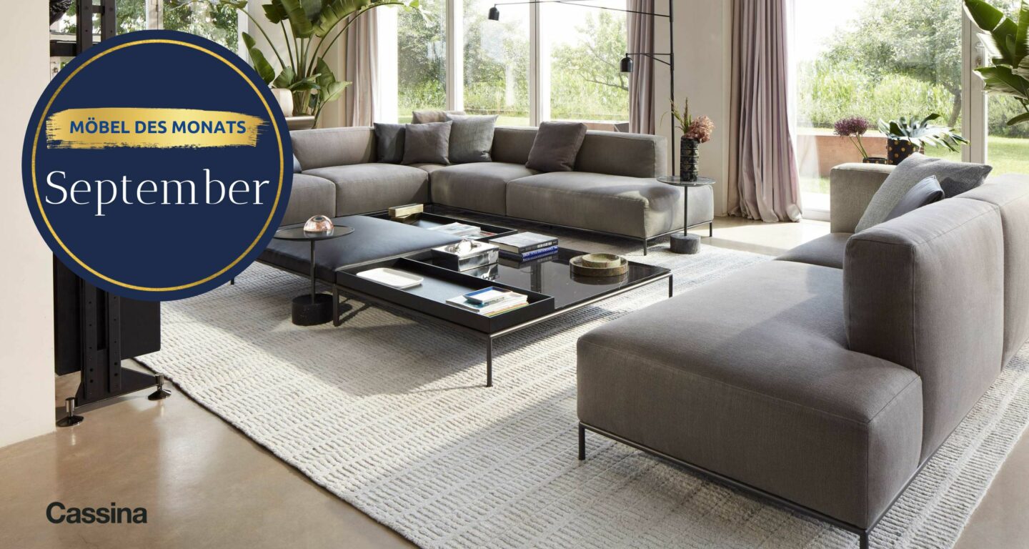 Modernes Wohnzimmerinterieur mit großem grauen Sektionssofa, schwarzem Couchtisch und bodentiefen Fenstern; rundes Schild mit der Aufschrift Möbel des Monats September im Möbelhaus Bad Homburg. Das Cassina-Logo ist sichtbar.