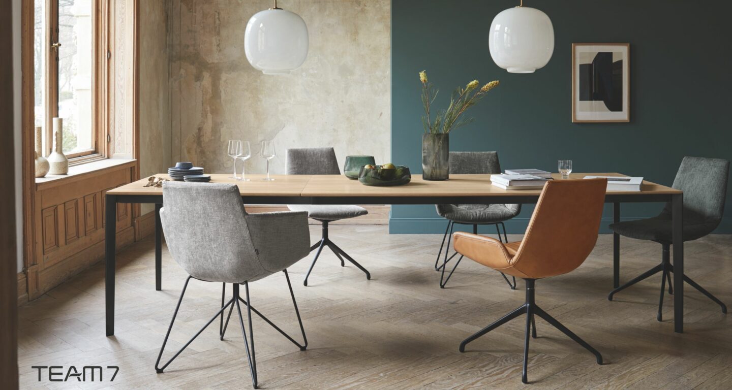 Modernes Esszimmer vom Möbelhaus Bad Homburg mit einem Holztisch, fünf verschiedenen Stühlen, Pendelleuchten, minimalistischem Dekor und einer Blumenvase auf einem Boden mit Fischgrätenmuster.