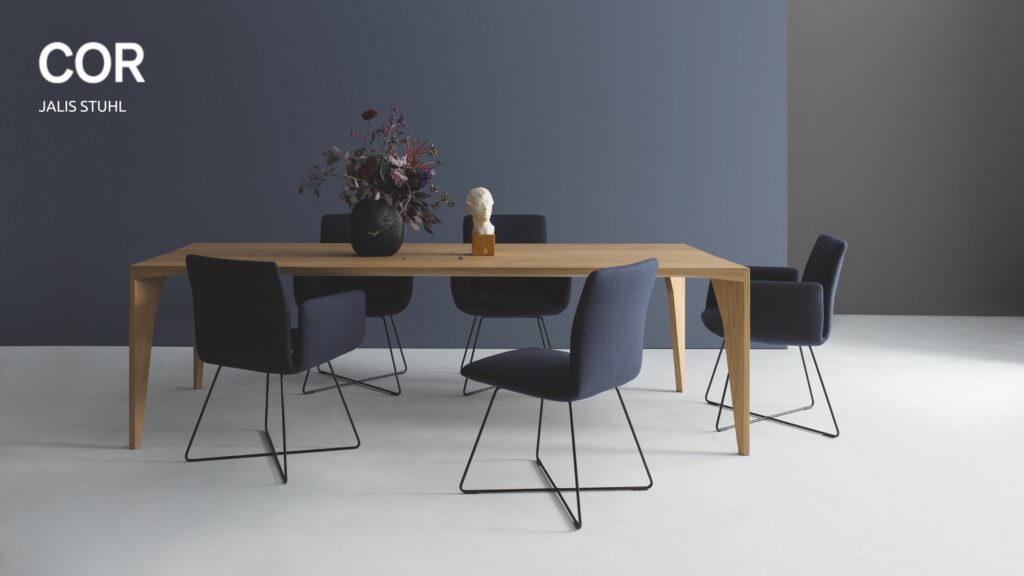 Ein moderner Esstisch aus Holz mit sechs dunkelblauen COR Jalis Stühlen, einer schwarzen Vase mit Blumen und einer kleinen weißen Büste darauf, vor einer blaugrauen Wand - jetzt in Aktion kaufen!.