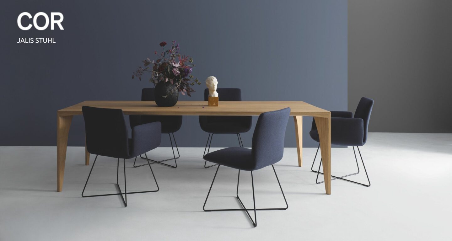 Ein moderner Esstisch aus Holz mit sechs dunkelblauen COR Jalis Stühlen, einer schwarzen Vase mit Blumen und einer kleinen weißen Büste darauf, vor einer blaugrauen Wand - jetzt in Aktion kaufen!.