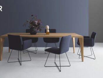 Ein moderner Esstisch aus Holz mit sechs dunkelblauen COR Jalis Stühlen, einer schwarzen Vase mit Blumen und einer kleinen weißen Büste darauf, vor einer blaugrauen Wand - jetzt in Aktion kaufen!.
