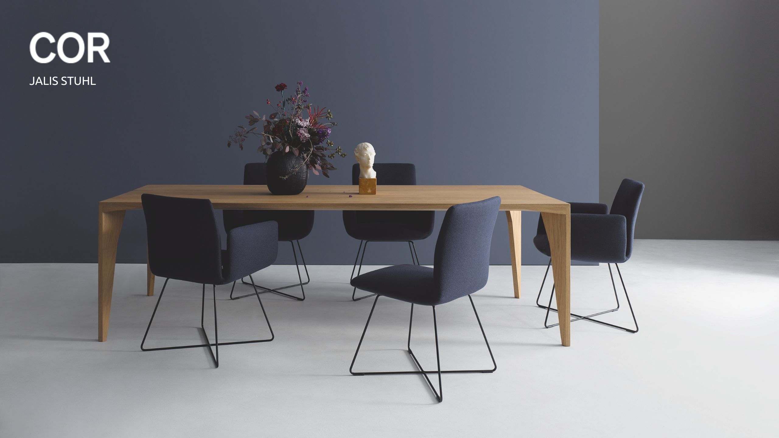 Ein moderner Esstisch aus Holz mit sechs dunkelblauen COR Jalis Stühlen, einer schwarzen Vase mit Blumen und einer kleinen weißen Büste darauf, vor einer blaugrauen Wand - jetzt in Aktion kaufen!.